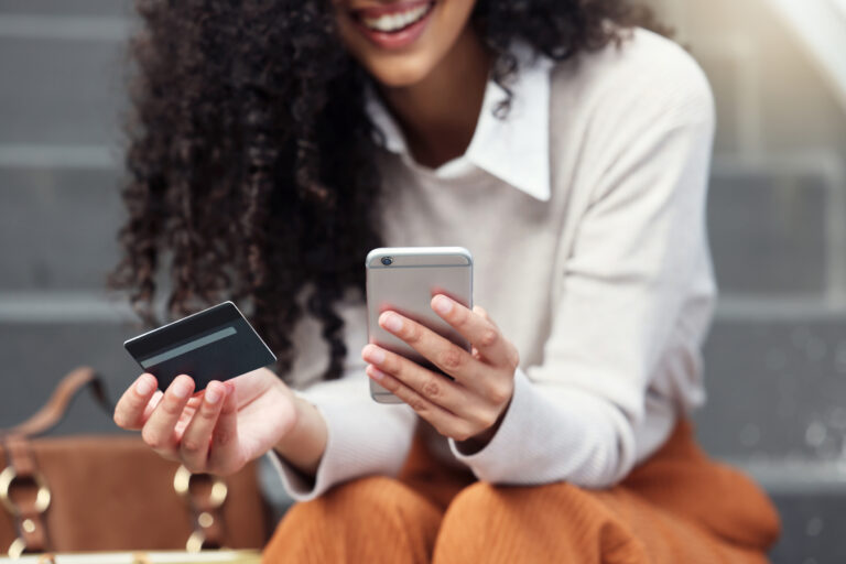 Mulher de camisa social sorrindo, segurando em uma mão um cartão de crédito e na outra um celular.