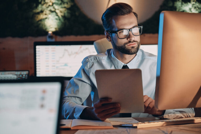 Um homem branco usando óculos se posiciona em frente a uma ela de computador, um tablet e mais outros computadores em um ambiente de escritório.