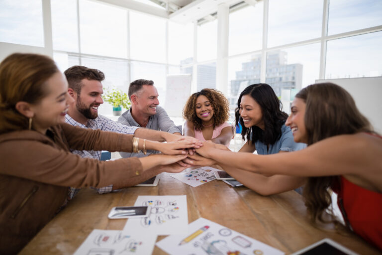 Seis pessoas sorridentes juntam as mão ao centro de uma mesa.